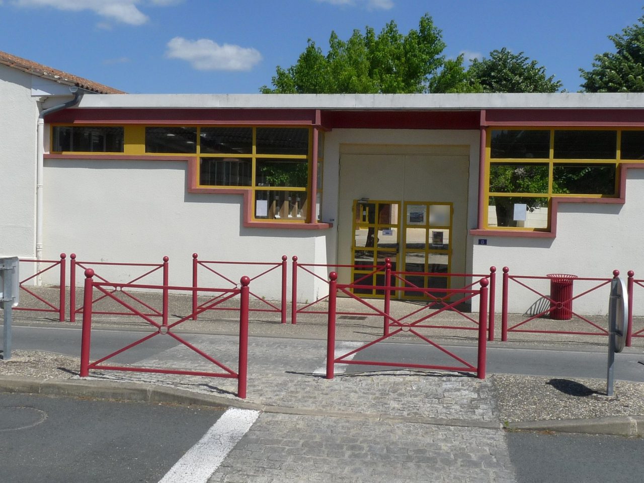 L'entrée de l'école côté rue : bâtiment blanc aux menuiseries jaunes, double porte vitrée, barrières rouges devant.